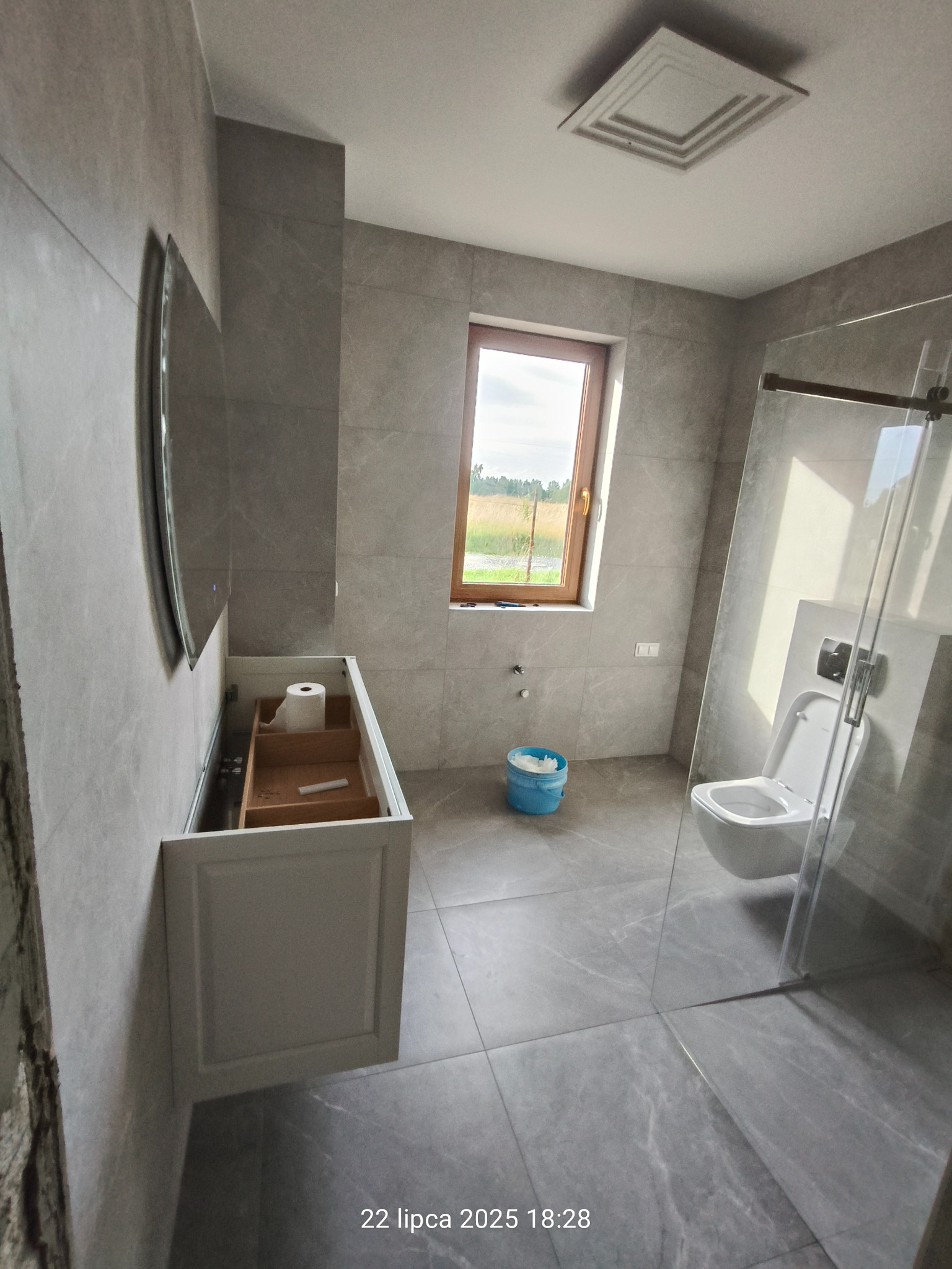 Nowoczesna łazienka z szarymi płytkami, kabiną prysznicową typu walk-in i drewnianym oknem. Umywalka w trakcie montażu, widoczny fragment lustra i toaleta.