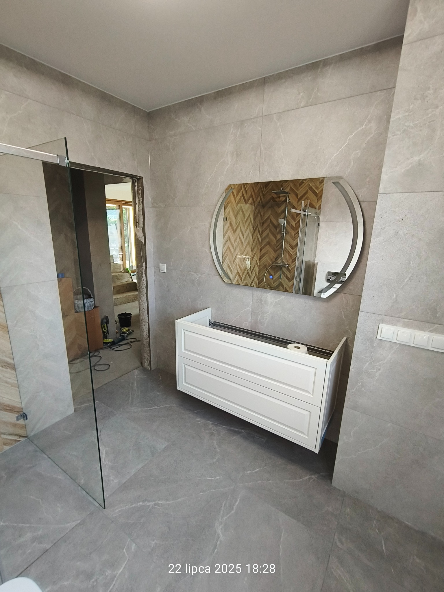 Łazienka w trakcie wykańczania: szare płytki, biała szafka pod umywalkę, lustro w owalnej ramie. Widoczna kabina prysznicowa i narzędzia w tle.
