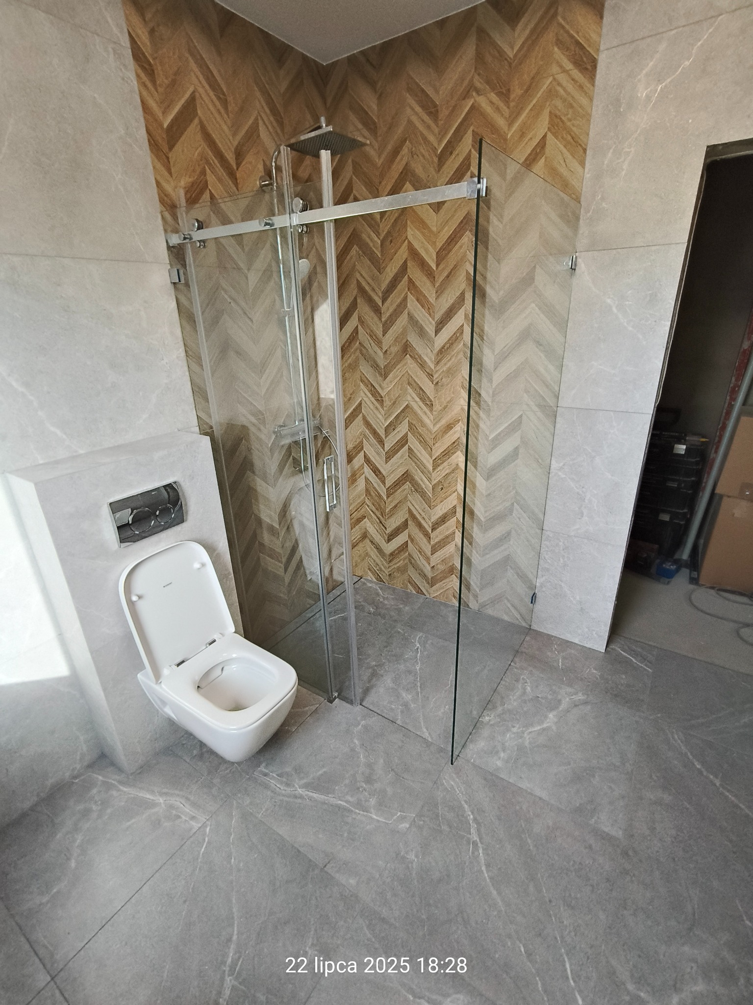 Nowoczesna łazienka po remoncie: kabina prysznicowa ze szklanymi drzwiami przesuwnymi, toaleta podwieszana i płytki imitujące marmur na podłodze i ścianach.