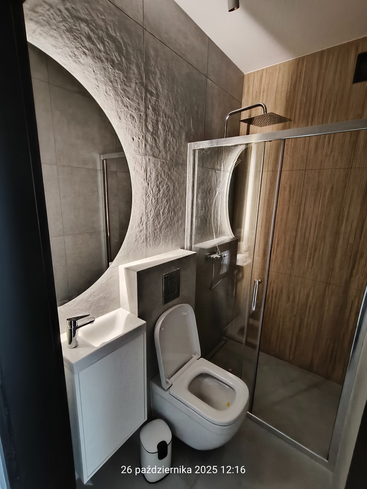 Nowoczesna łazienka z umywalką nablatową, toaletą i kabiną prysznicową. Ściany wykończone płytkami imitującymi beton i drewno. Lustro w kształcie łuku.