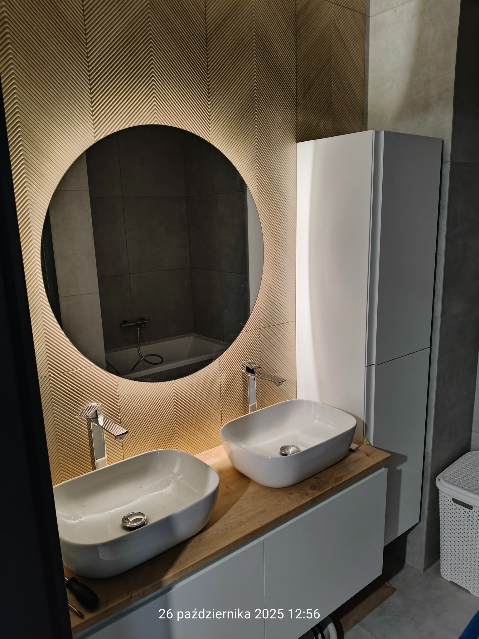 Nowoczesna łazienka z dwiema umywalkami nablatowymi, okrągłym lustrem z podświetleniem LED i dekoracyjną ścianą z wzorem jodełki. Minimalistyczny design i eleganckie wykończenie.