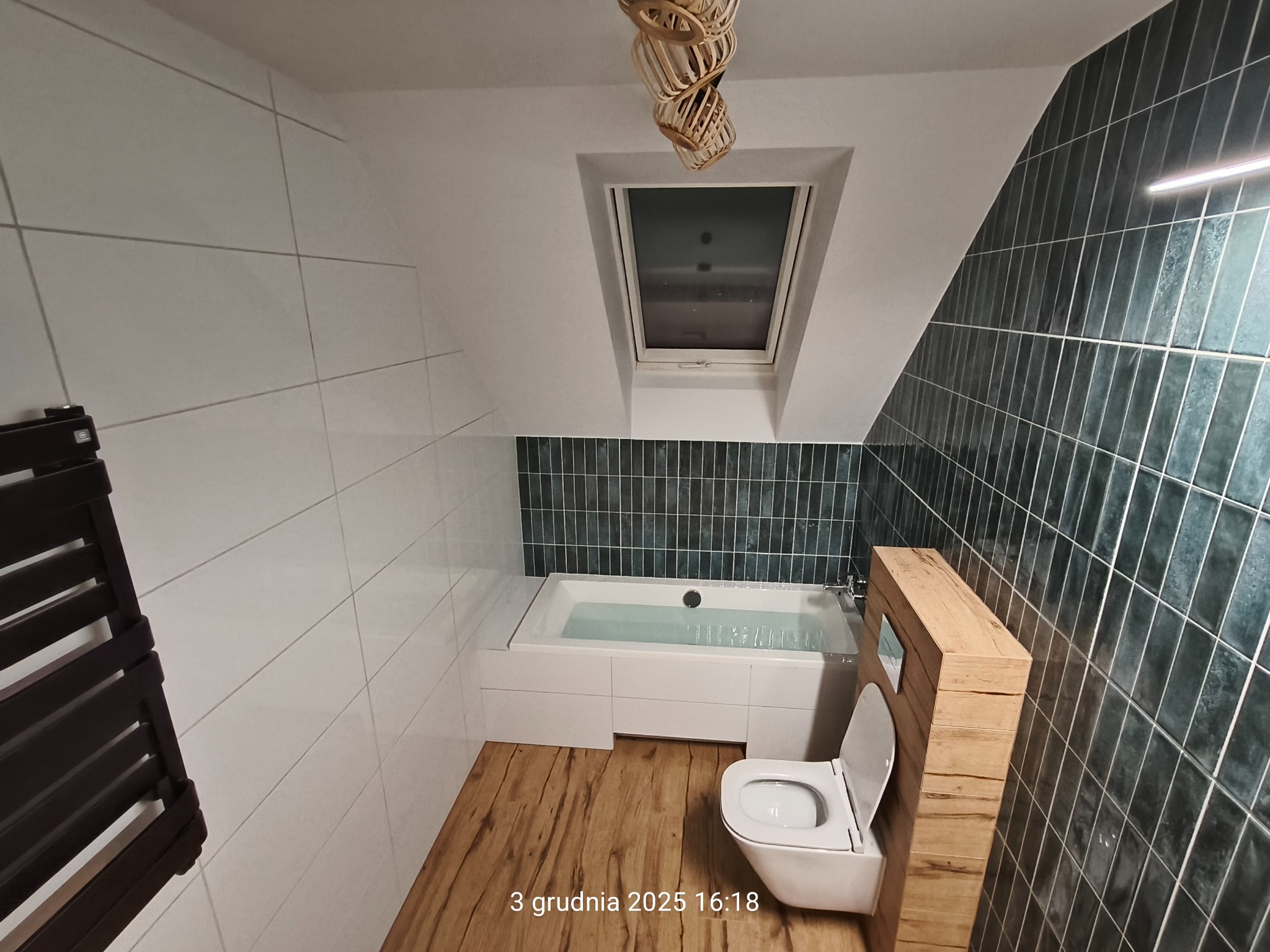 Nowoczesna łazienka na poddaszu z wanną, toaletą w zabudowie drewnianej i grzejnikiem drabinkowym. Uwagę przykuwają ciemne, pionowe płytki na ścianie.