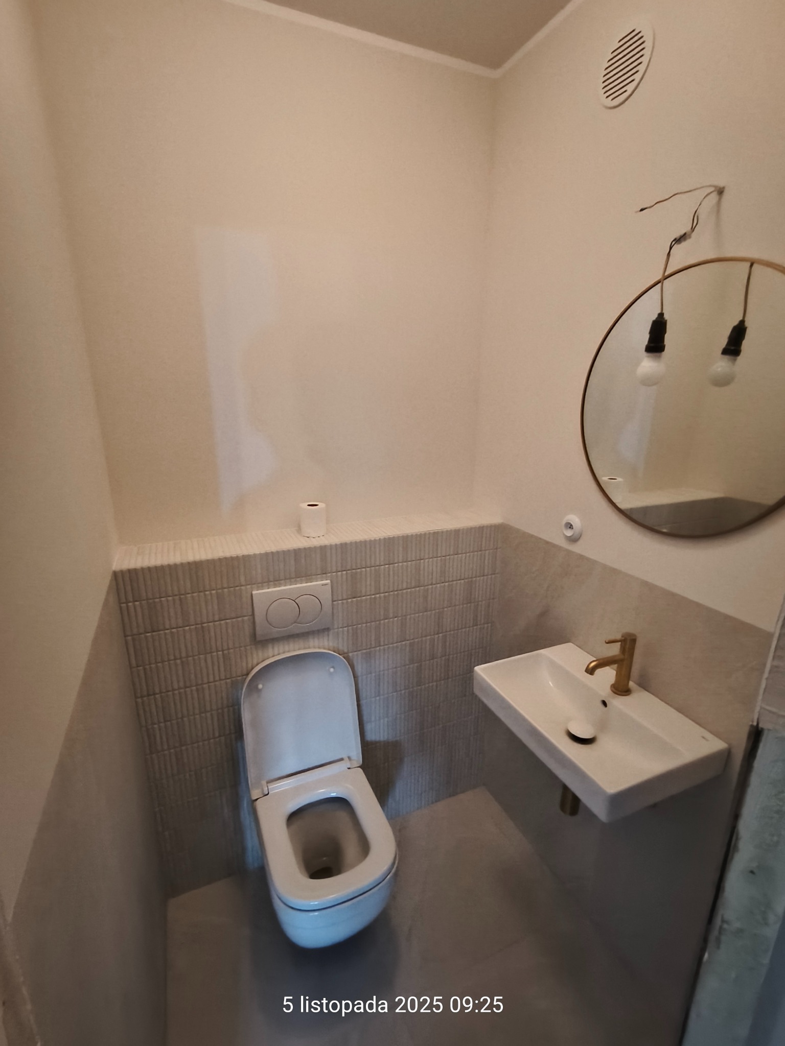 Nowoczesna, jasna łazienka po remoncie: wisząca toaleta, umywalka z złotą baterią, okrągłe lustro z żarówkami. Minimalistyczny design, płytki imitujące cegłę.