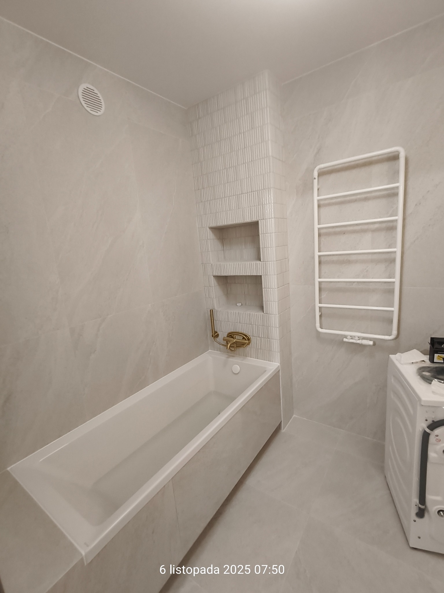 Jasna łazienka po remoncie: wanna z złotą armaturą, grzejnik drabinkowy, płytki imitujące marmur i wnęki w ścianie wyłożone drobną mozaiką. Widoczna pralka.