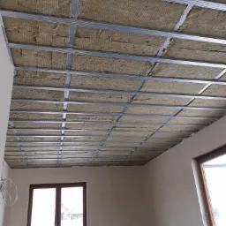 Sufit podwieszany z metalową konstrukcją wypełnioną wełną mineralną, widok z dołu w trakcie montażu w jasnym pomieszczeniu z oknami.