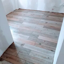 Ułożona podłoga z płytek imitujących drewno w odcieniach szarości i beżu, widoczne niedokończone ściany i wystający kabel elektryczny.