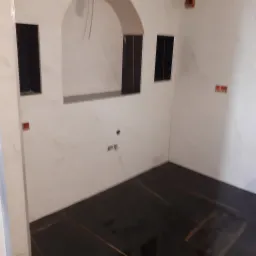 Wnętrze łazienki w trakcie remontu, wyłożone białymi płytkami imitującymi marmur z ciemnymi wstawkami w niszach i na podłodze, widoczne wystające przewody elektryczne.