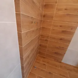 Wykończona kabina prysznicowa z brązowymi płytkami imitującymi drewno, jasnoszare ściany, widoczne mocowania na ścianie i w podłodze.