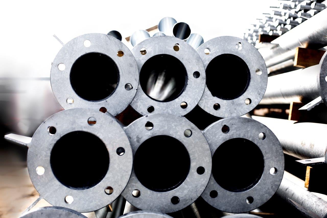 Stos ocynkowanych stalowych kołnierzy z otworami na śruby, ułożonych na palecie z drewnianymi przekładkami, widoczne również rury w tle.