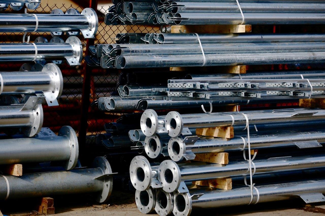 Stos ocynkowanych, stalowych słupów z kołnierzami, zabezpieczonych taśmą, ułożonych na drewnianych legarach na placu budowy.