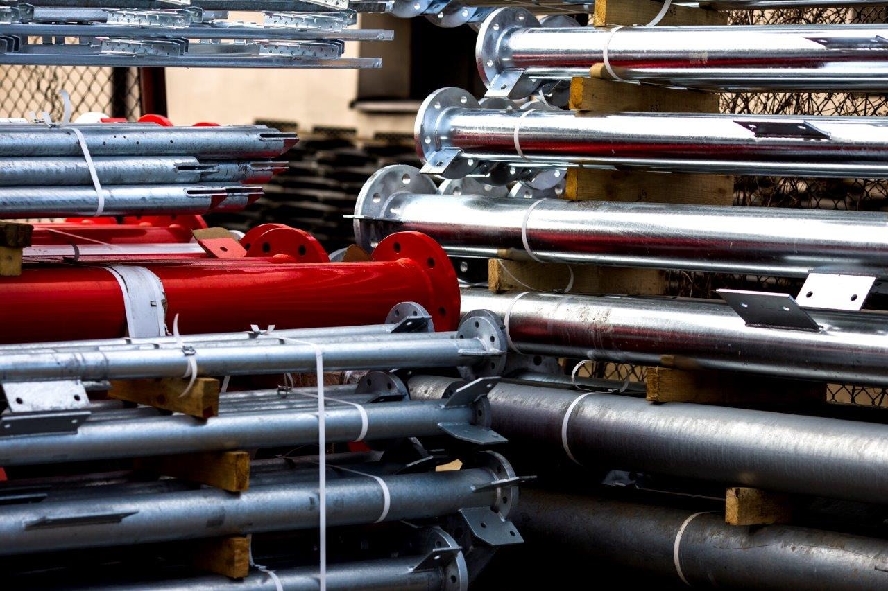 Stalowe rury i słupy o różnych średnicach, niektóre pomalowane na czerwono, składowane na drewnianych paletach, zabezpieczone białymi opaskami zaciskowymi.