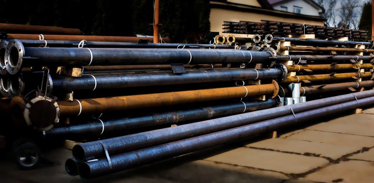 Stalowe rury z kołnierzami ułożone na drewnianych legarach, niektóre z widoczną rdzą, zabezpieczone białymi taśmami.