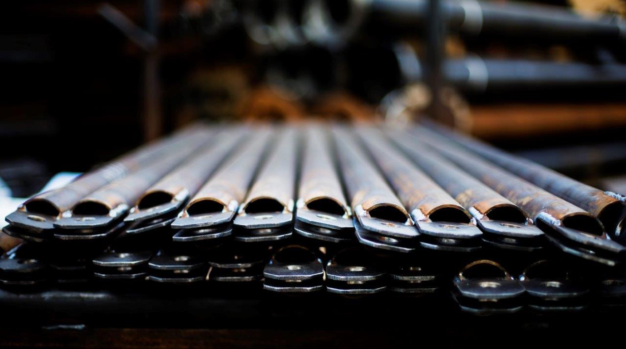 Rząd surowych, metalowych rur z przyspawanymi płaskownikami, widoczne ślady spawania i rdzy, ułożone równolegle na ciemnym tle.