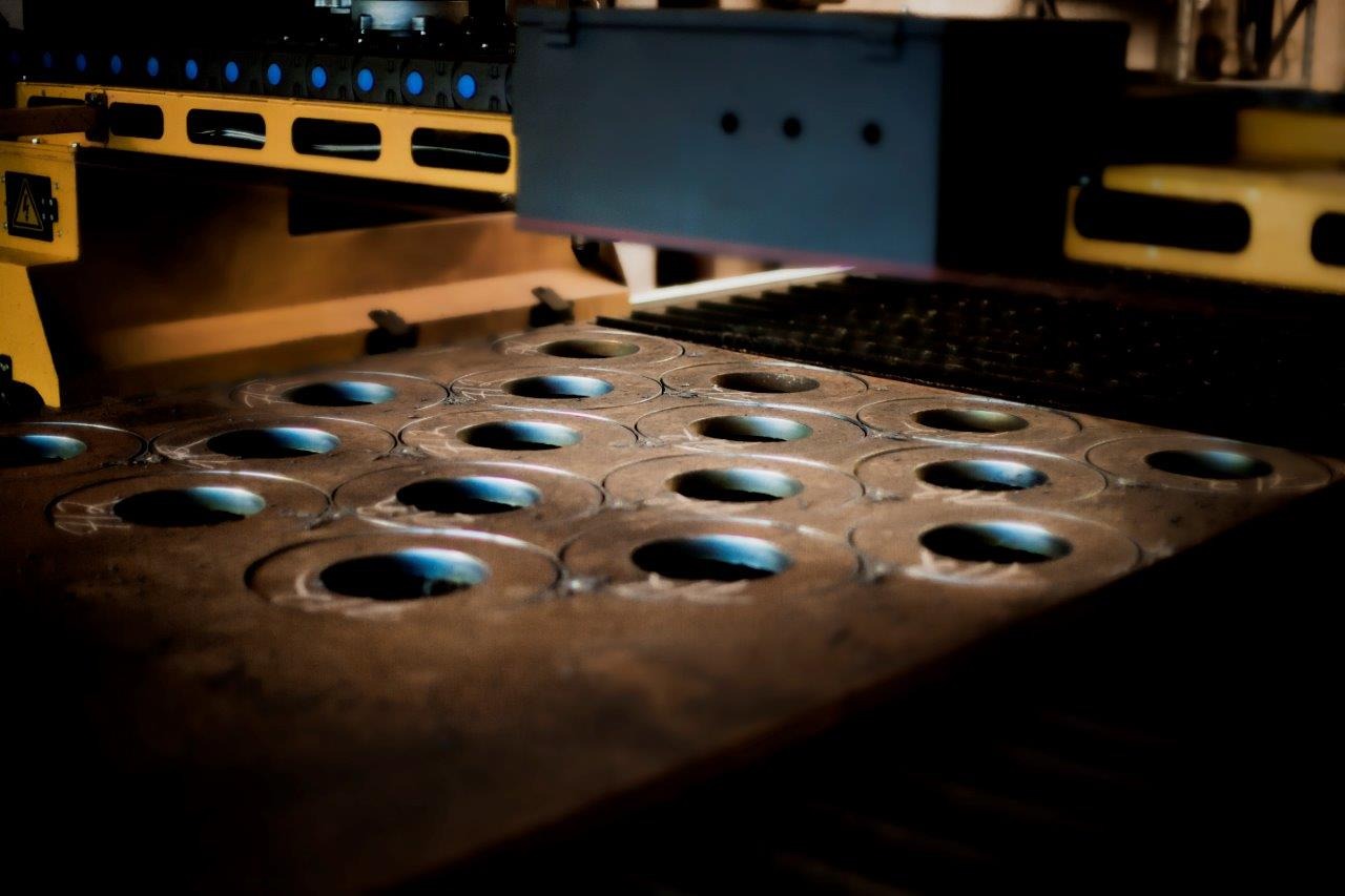 Proces wycinania otworów w metalowej płycie za pomocą maszyny CNC; widoczne liczne okrągłe otwory w brązowawej płycie, żółte elementy maszyny w tle, precyzyjne wycinanie.