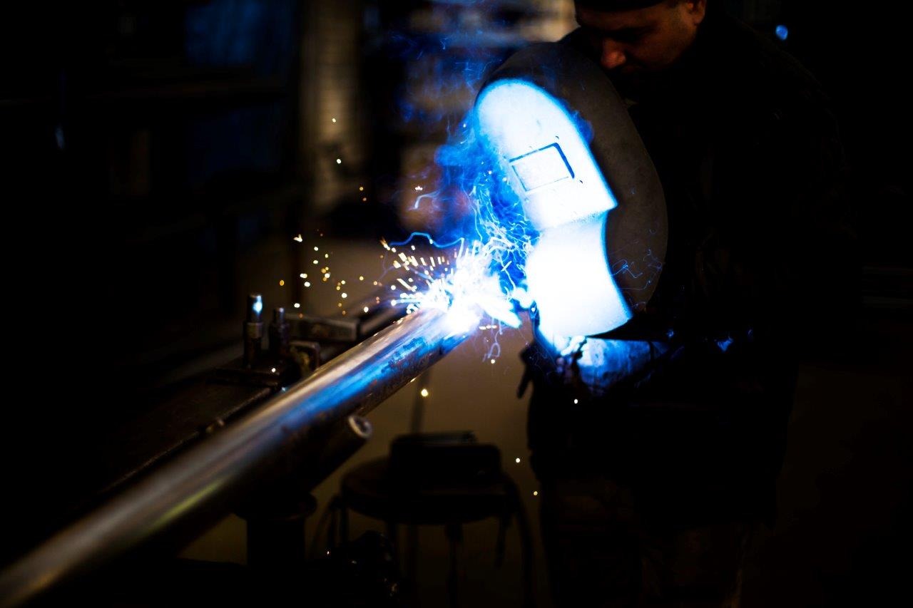 Spawanie rury przez spawacza w masce, iskry i niebieskie światło łuku elektrycznego, ciemne tło.