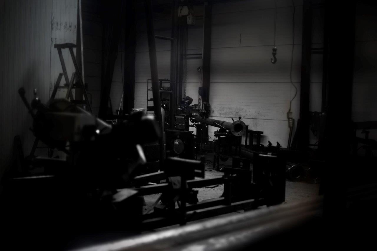 Ciemne wnętrze warsztatu z wyposażeniem do obróbki metalu, w tle drabina i hak wiszący z sufitu.