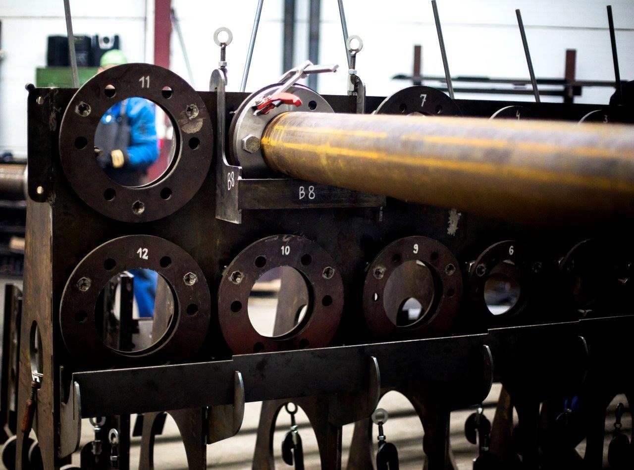 Metalowa konstrukcja z otworami oznaczonymi numerami, przez jeden z otworów przechodzi rura. W tle widoczna osoba w niebieskim kombinezonie.