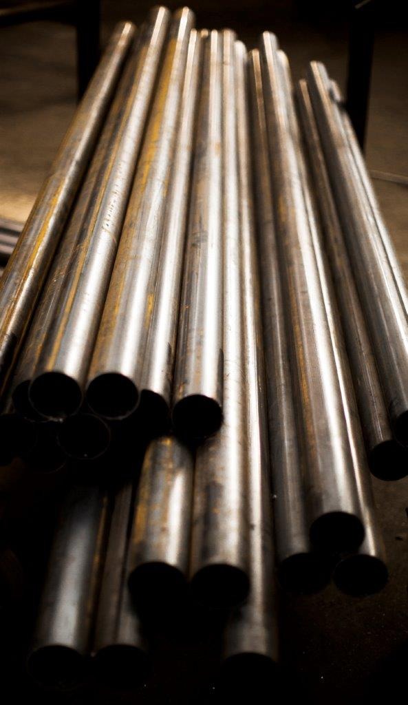 Stos srebrzystych, metalowych rur o widocznych śladach korozji, ułożonych równolegle w ciemnym otoczeniu warsztatowym, z widocznymi przekrojami.