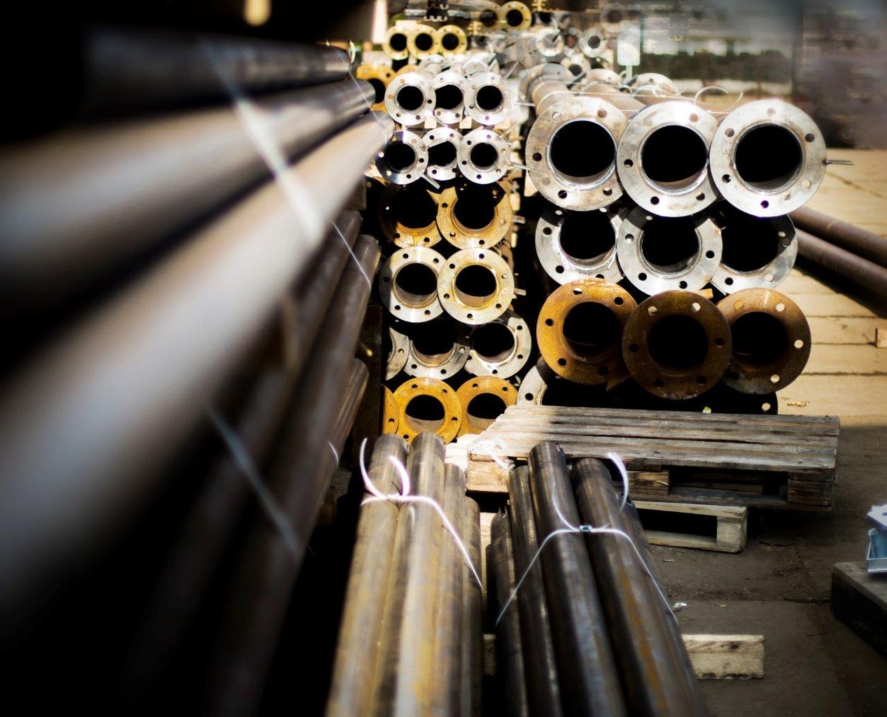 Stalowe rury i kołnierze w magazynie w Warszawie, ułożone w stosy na paletach, widoczne ślady korozji na niektórych elementach.