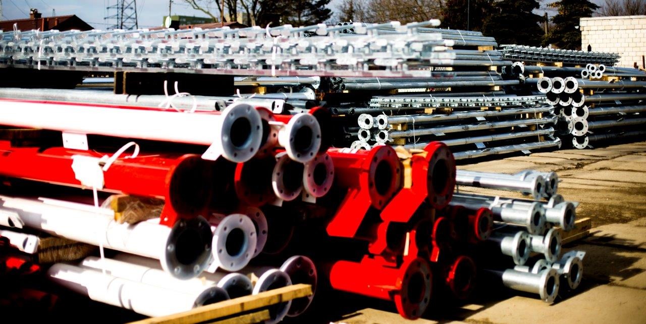 Stosy rur stalowych z kołnierzami w kolorach srebrnym, czerwonym i białym, ułożone na zewnątrz w magazynie lub na placu budowy, widoczne elementy konstrukcyjne w tle.