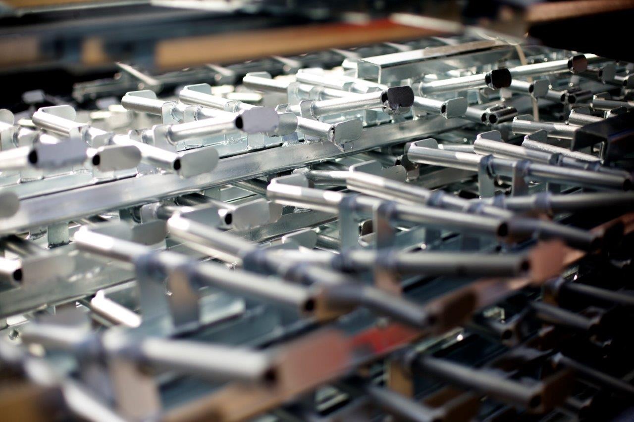 Szereg ocynkowanych, powtarzalnych elementów metalowych, prawdopodobnie wsporników lub uchwytów, ułożonych warstwami, tworzących uporządkowany wzór z elementami w kolorze srebrnym i czarnym.