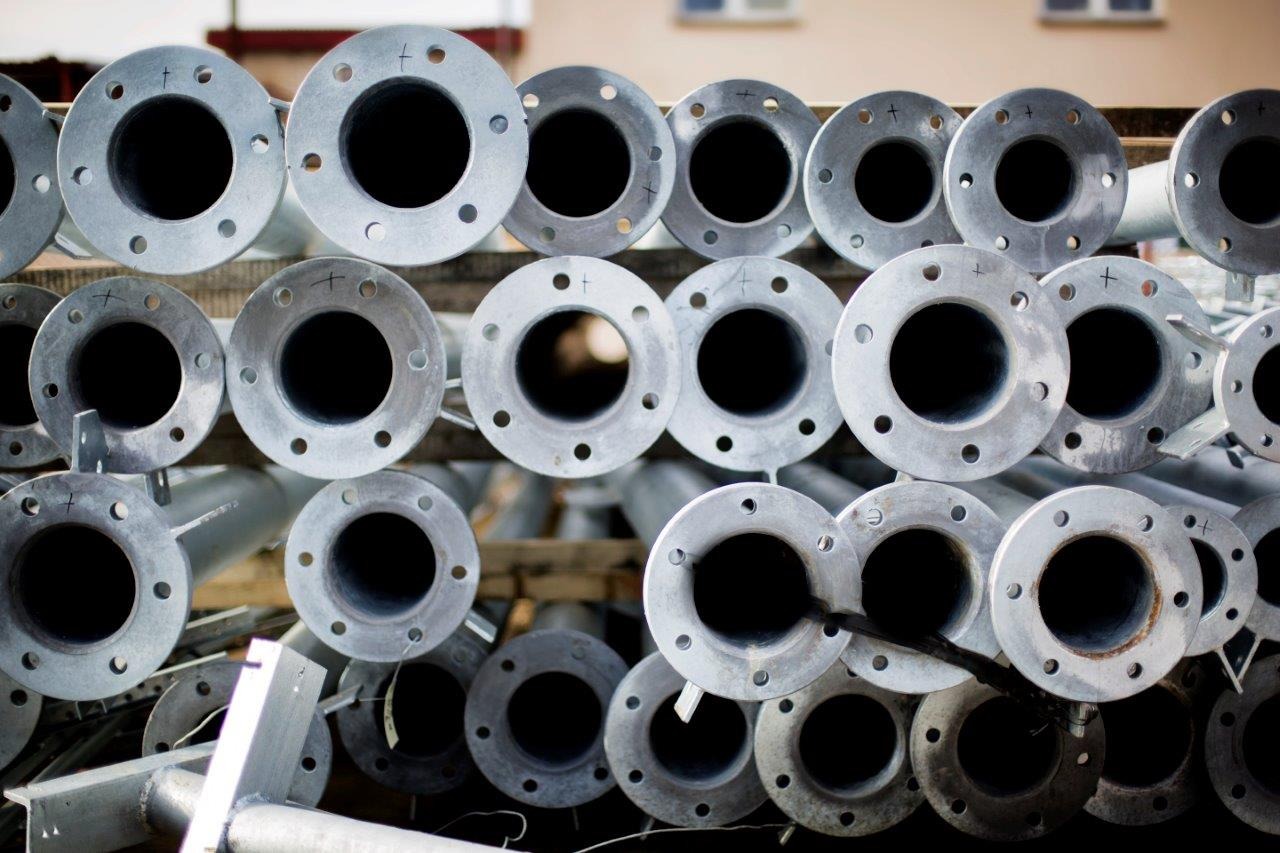 Stos ocynkowanych stalowych rur z kołnierzami, ułożonych na drewnianej palecie, widoczne znaki montażowe na niektórych elementach.