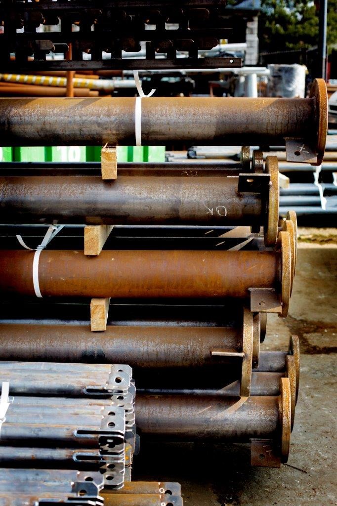 Stos rdzewiejących stalowych rur z kołnierzami, zabezpieczonych drewnianymi przekładkami i białymi opaskami zaciskowymi, widoczne elementy konstrukcyjne w tle.