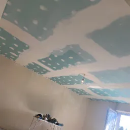 Niedokończony sufit z płyt kartonowo-gipsowych z widocznymi łączeniami szpachlowanymi, narzędzia na stole warsztatowym, zabezpieczona folią okno oraz widoczne ślady szpachli na ścianach.