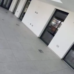 Jasnoszara podłoga z kwadratowych płytek, widoczne czarne ramy okien i drzwi, ściany pomalowane na biało, z widocznymi puszkami elektrycznymi i elementami wentylacji pod sufitem.