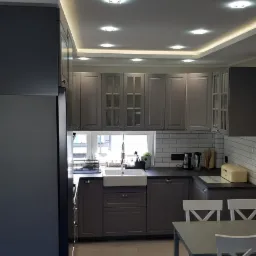 Nowoczesna kuchnia w odcieniach szarości z białymi akcentami, oświetleniem sufitowym LED i białym ceramicznym zlewem farmerskim pod oknem.