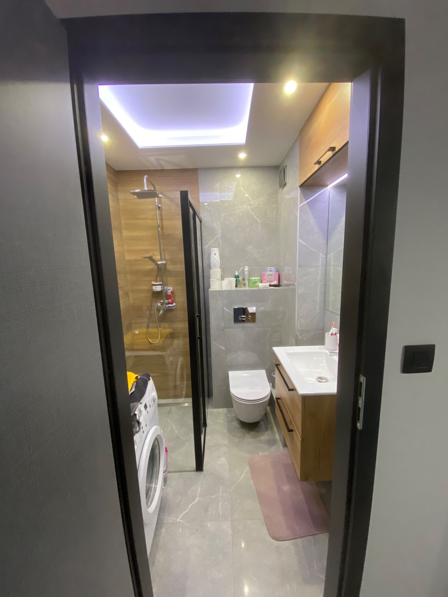 Nowoczesna łazienka z kabiną prysznicową typu walk-in, umywalką z szafką z jasnego drewna, podświetlanym lustrem i pralką widoczną w tle, sfotografowana zza futryny drzwi.