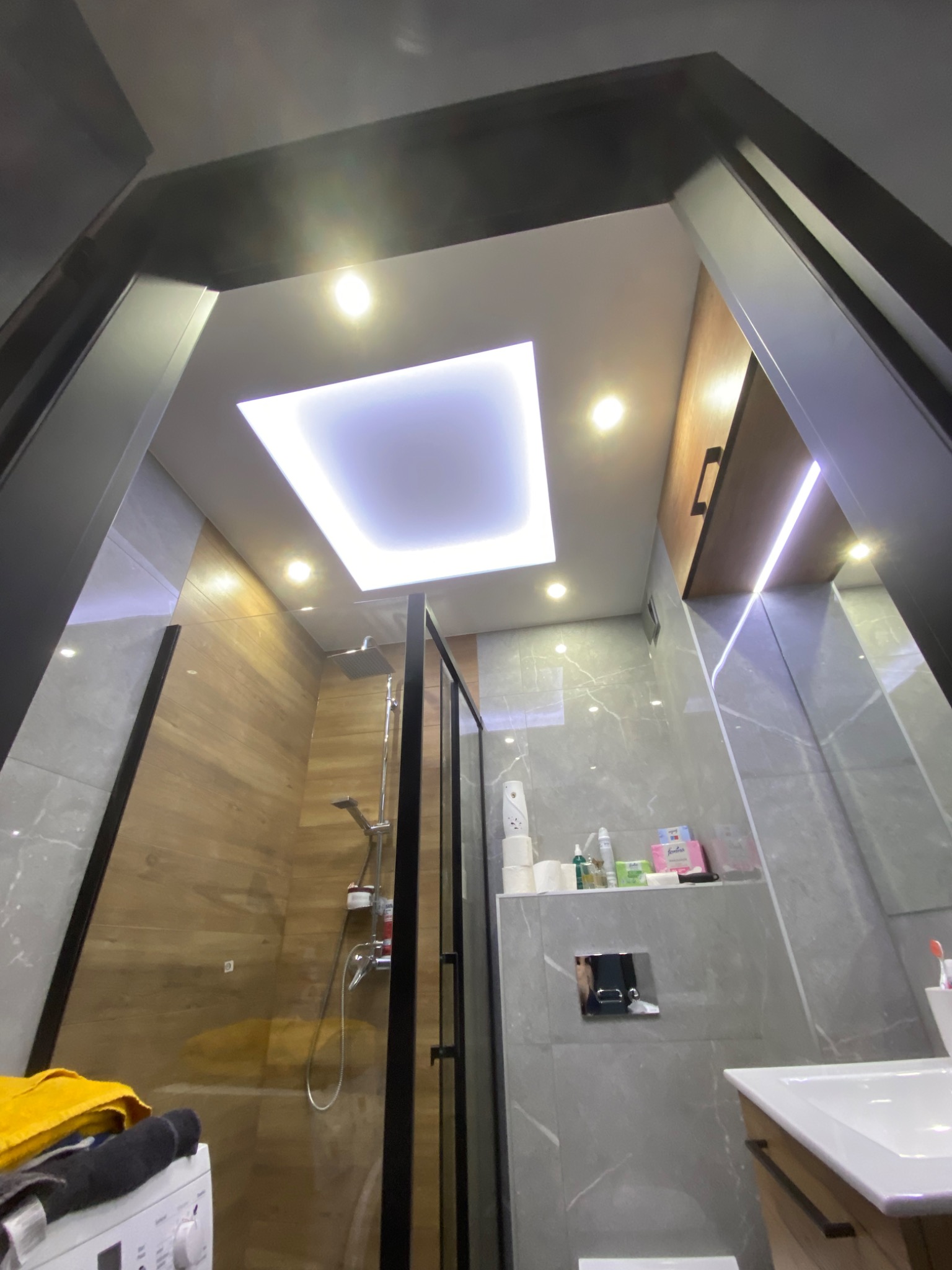 Nowoczesna łazienka z podświetlanym sufitem, kabiną prysznicową typu walk-in z czarnymi ramami, płytkami imitującymi drewno i marmur, oraz widoczną pralką z żółtym ręcznikiem.