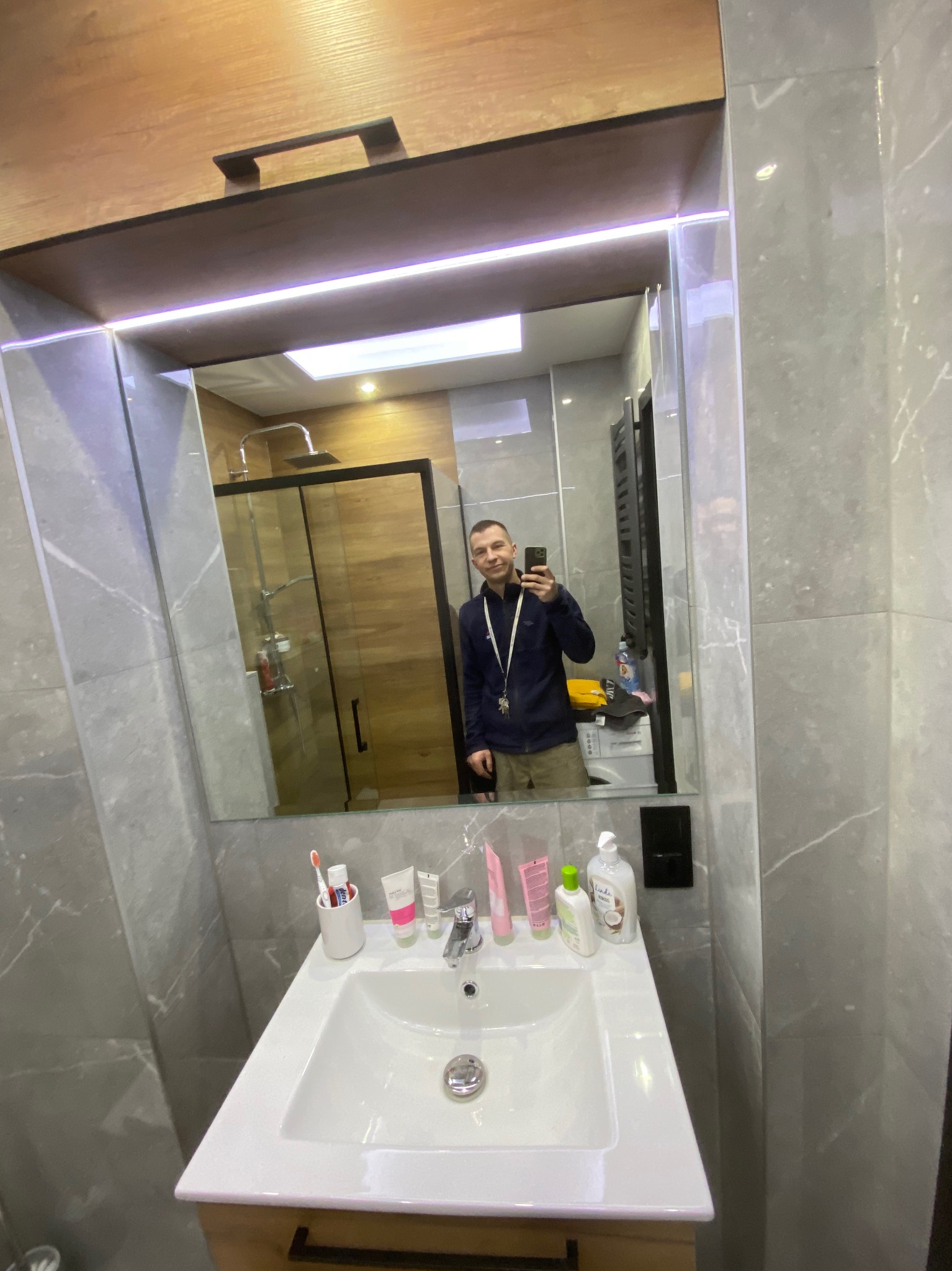 Jasna łazienka z szarymi marmurkowymi płytkami, kabiną prysznicową typu walk-in i białym blatem umywalki, widoczna w odbiciu lustra osoba robiąca zdjęcie.