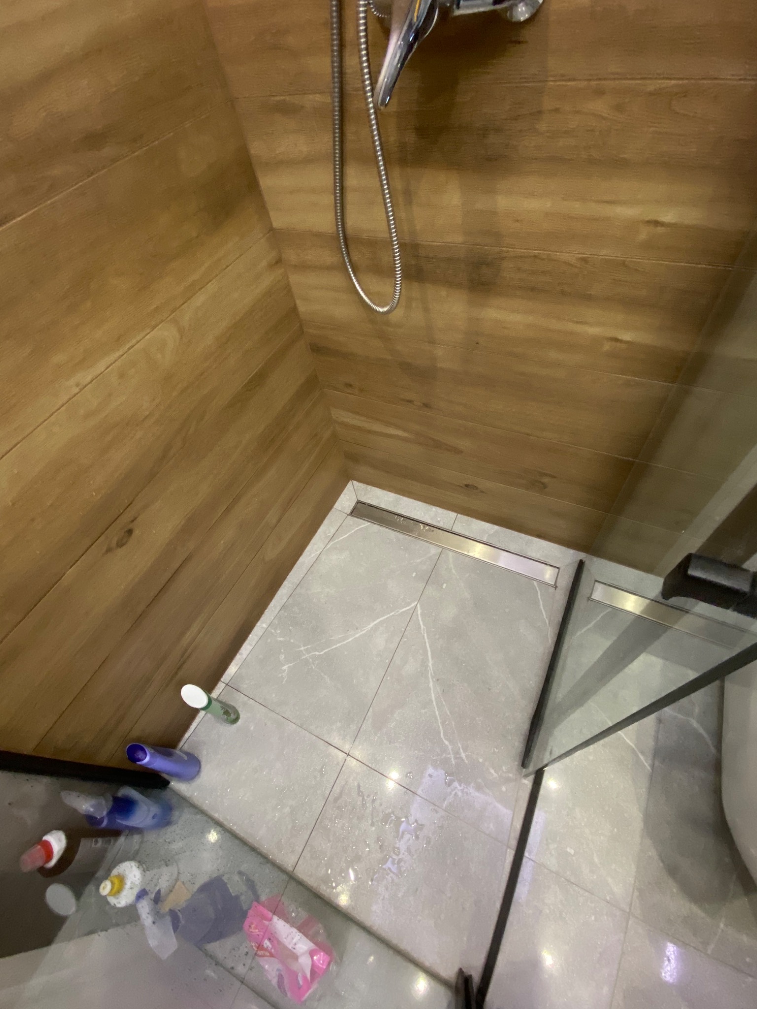 Szczegółowe ujęcie kabiny prysznicowej z odpływem liniowym, wyłożonej płytkami imitującymi drewno na ścianach i szarymi płytkami z marmurowym wzorem na podłodze, widoczne kosmetyki na podłodze.