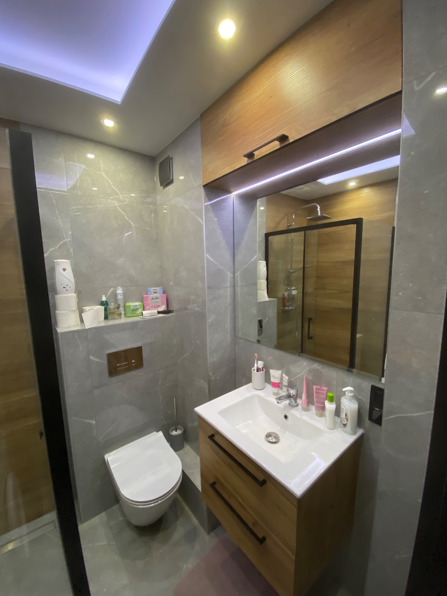 Nowoczesna łazienka w szarościach z drewnianymi akcentami: umywalka z szafką, wisząca toaleta i kabina prysznicowa z czarnymi ramami widoczna w lustrze.