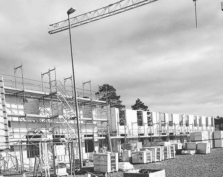 Budowa domu z prefabrykatów: widok na plac budowy z rusztowaniami i dźwigiem w tle, monochromatyczne ujęcie.