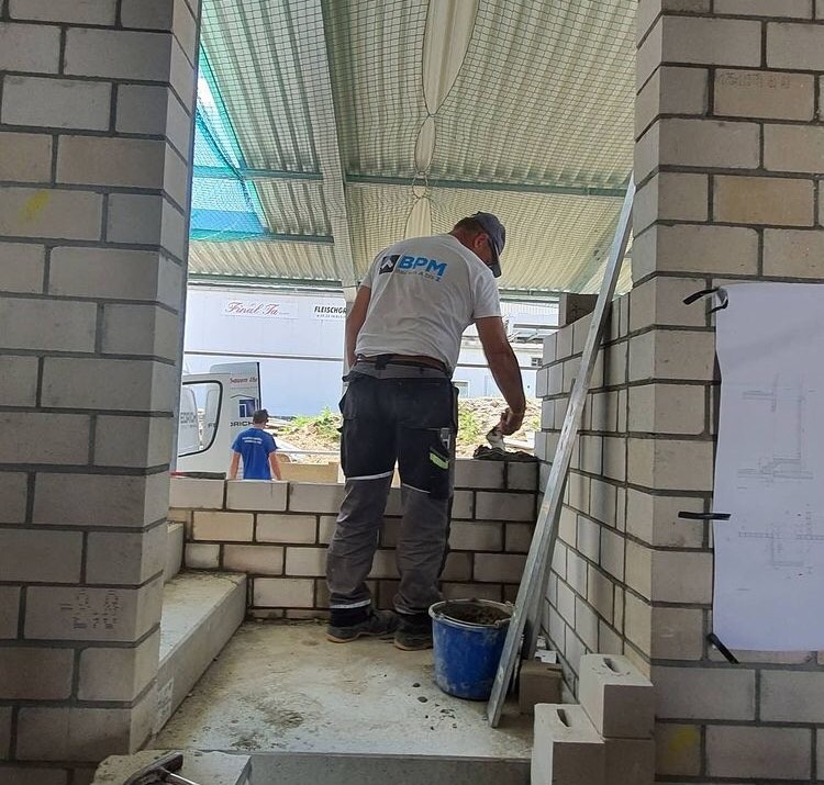 Murarz w trakcie wznoszenia ściany z jasnych cegieł; widoczny plan budynku przytwierdzony do ściany, wiadro z zaprawą oraz pracownik w tle.