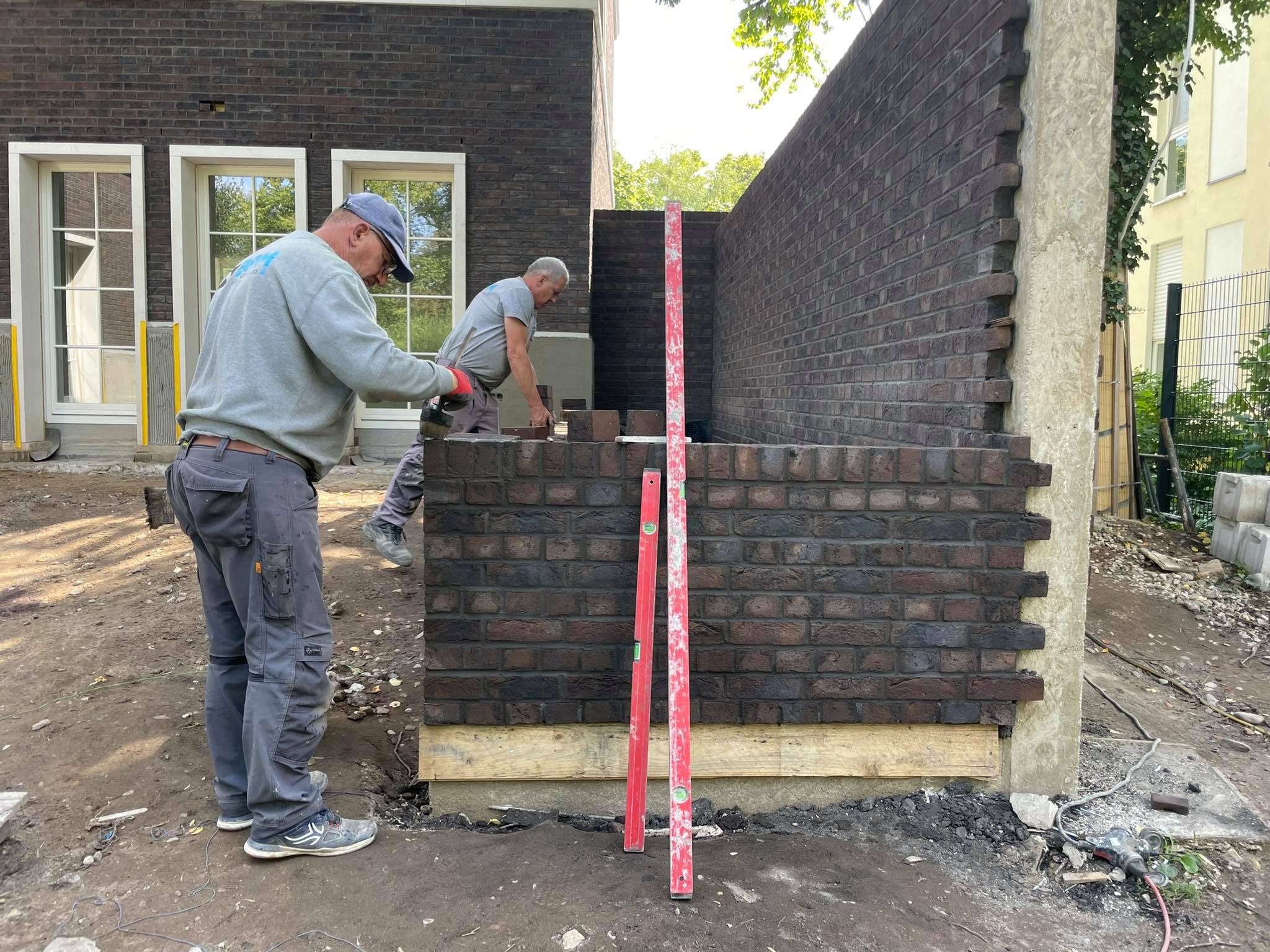 Dwóch murarzy w trakcie wznoszenia ściany z ciemnej cegły na zewnątrz budynku, widoczne poziomice i narzędzia.