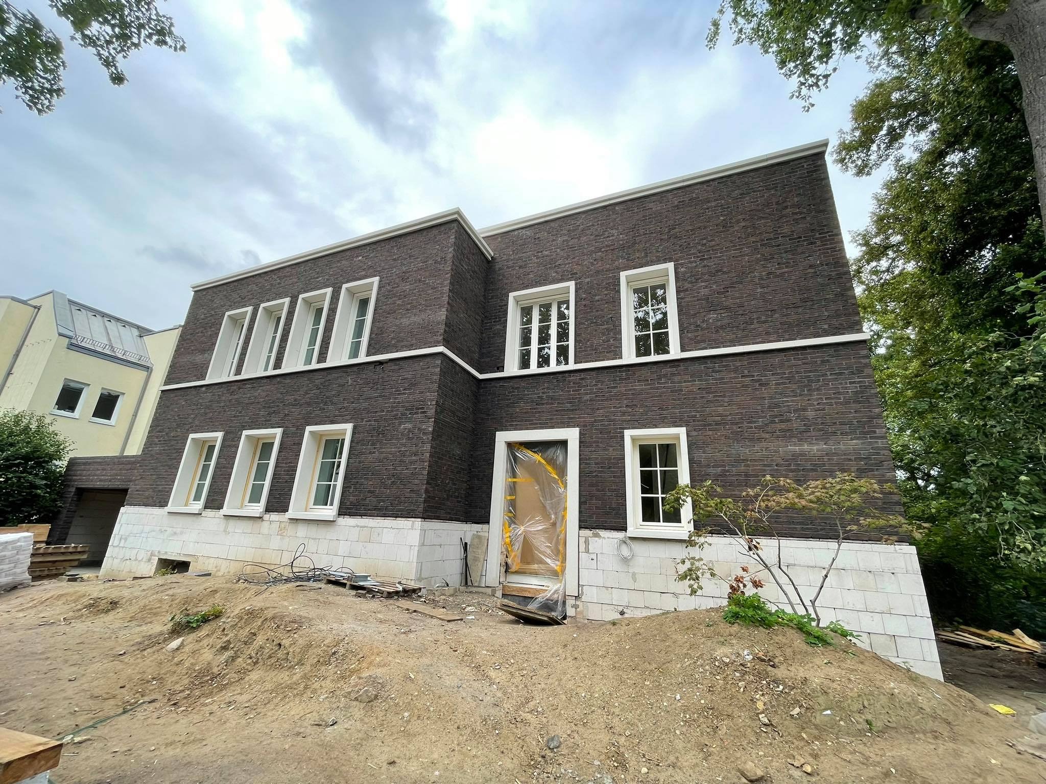 Nowoczesny, dwukondygnacyjny dom z ciemnej cegły klinkierowej z białymi obramowaniami okien i cokołem z bloczków. Wejście zabezpieczone folią ochronną. Teren wokół domu w trakcie prac ziemnych.