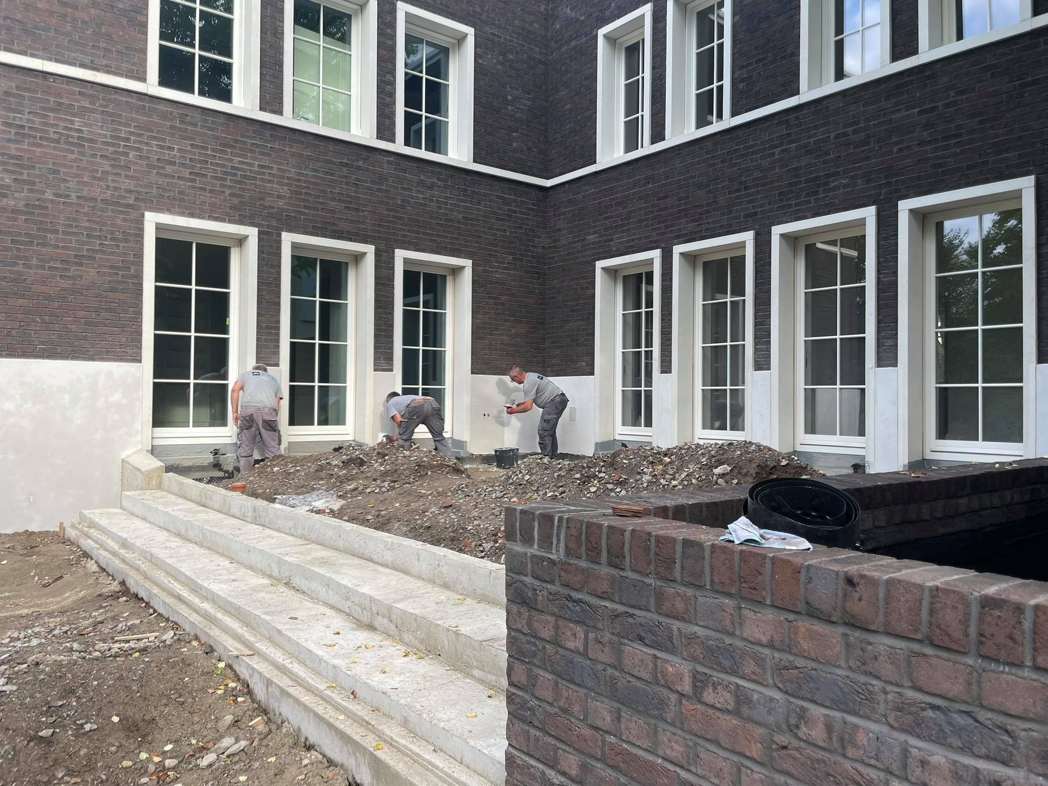 Trzech pracowników tynkuje elewację budynku z ciemnej cegły z białymi oknami, widoczne betonowe schody i fragment muru z cegły klinkierowej w pierwszym planie.
