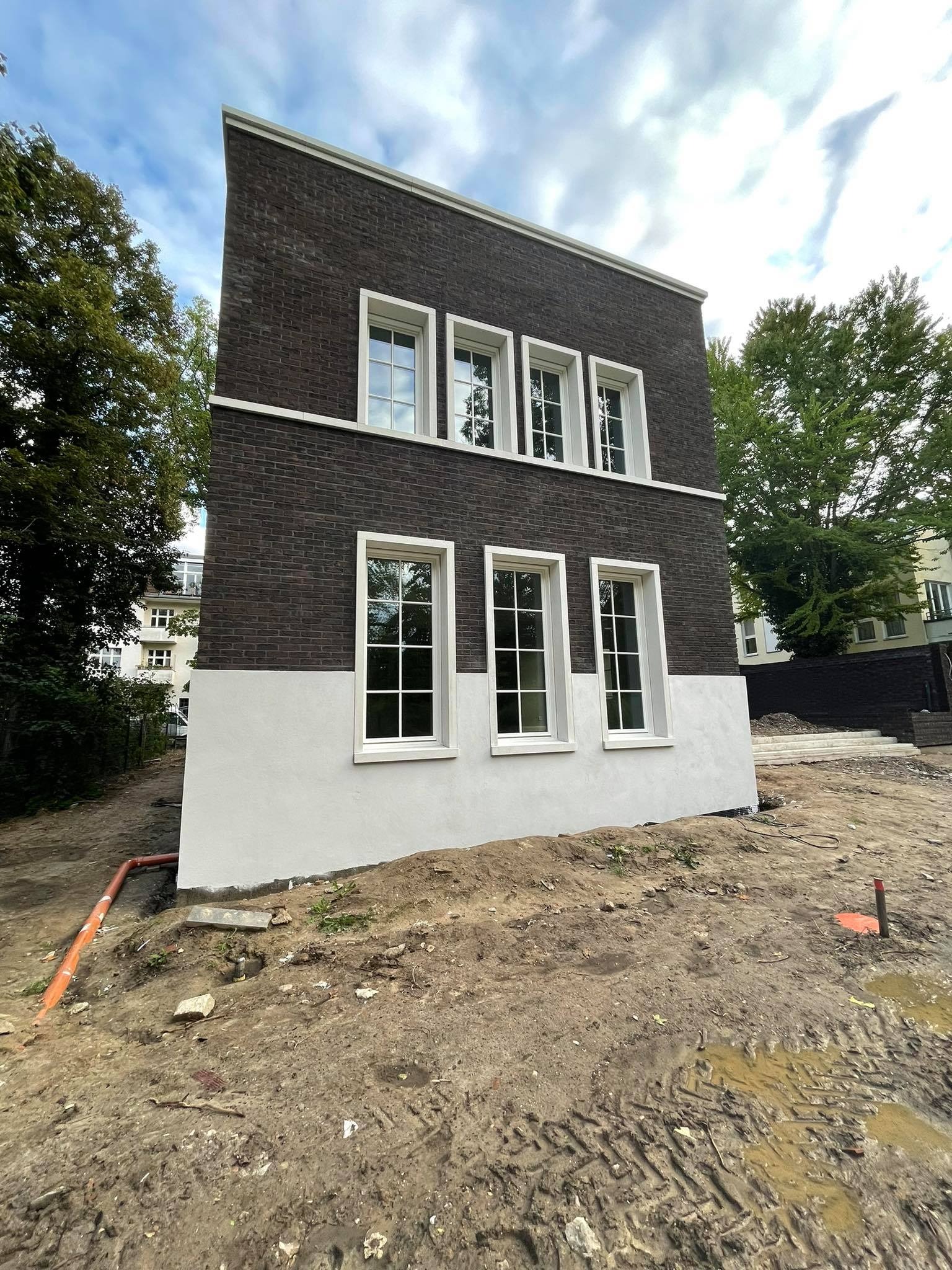 Surowy stan elewacji dwukondygnacyjnego budynku z ciemnej cegły z białymi ramami okiennymi i pasem tynku, widoczne ślady prac ziemnych wokół budynku, pomarańczowa rura w ziemi.