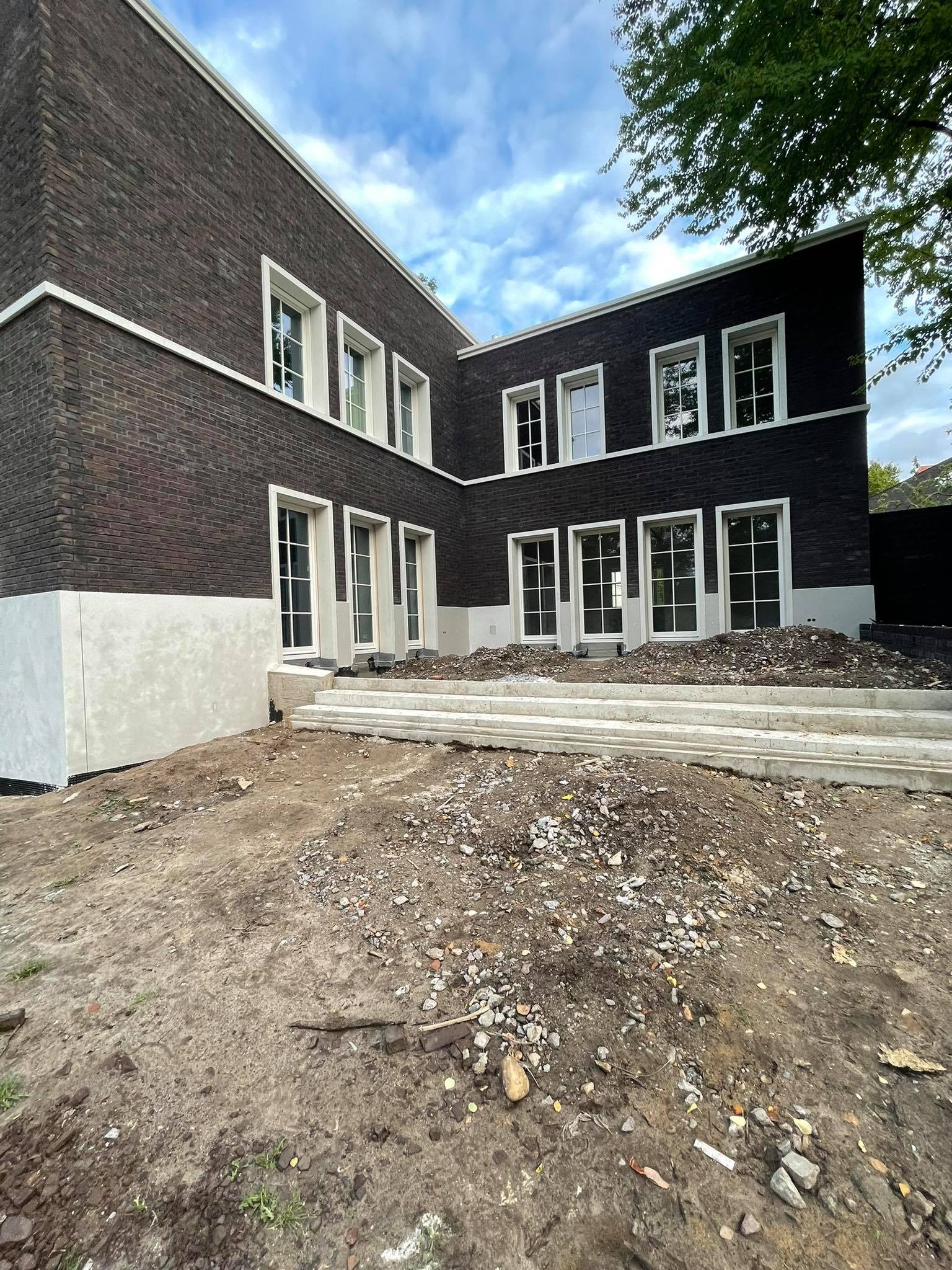 Surowy stan budowy domu z ciemnej cegły z białymi ramami okiennymi i betonowymi schodami wejściowymi, widoczne prace wykończeniowe wokół budynku.