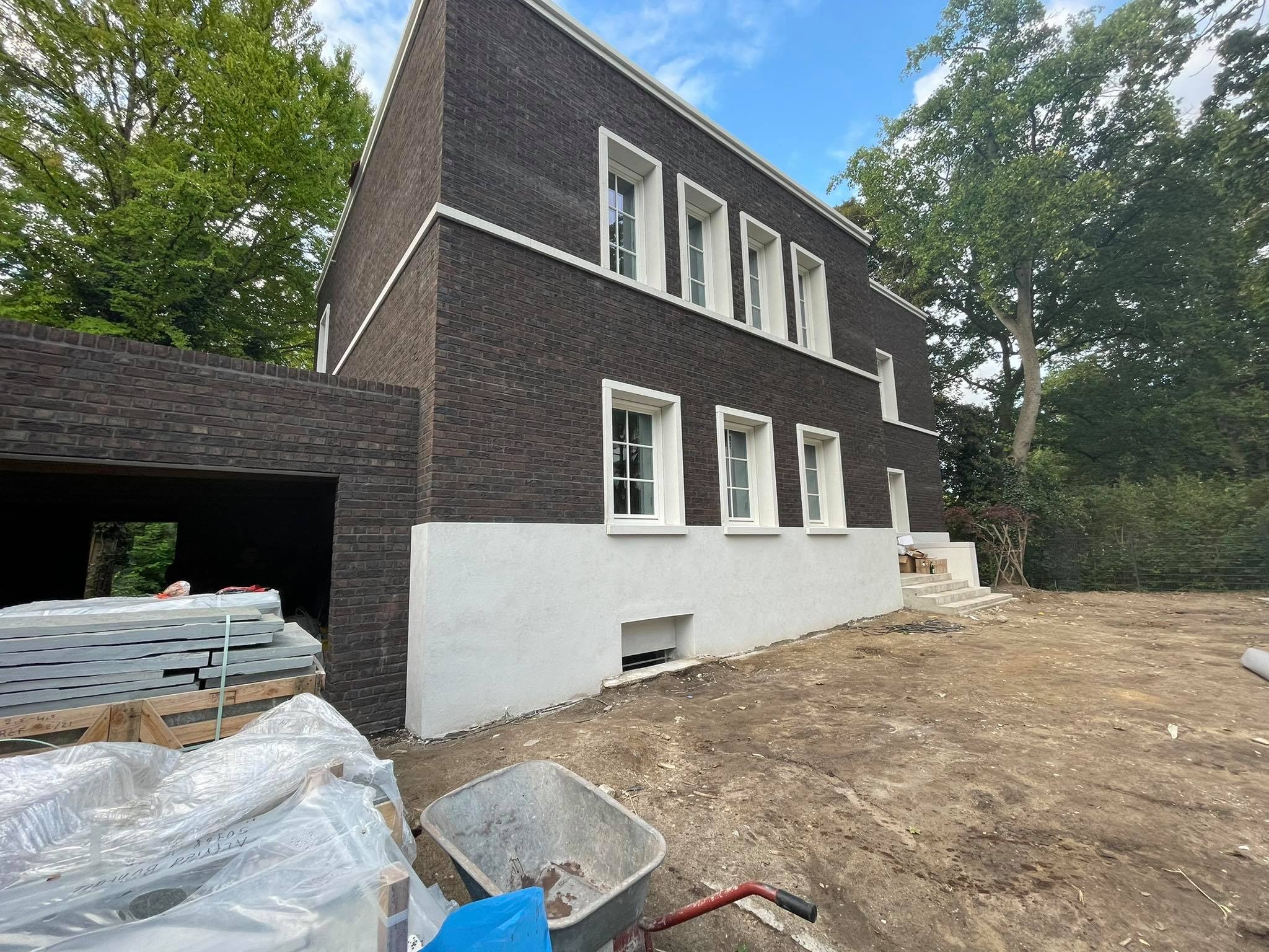 Nowoczesny dom murowany z ciemnej cegły z białymi ramami okiennymi i wykończeniem, widok z zewnątrz na etapie prac wykończeniowych, z widocznymi materiałami budowlanymi i taczką na pierwszym planie.