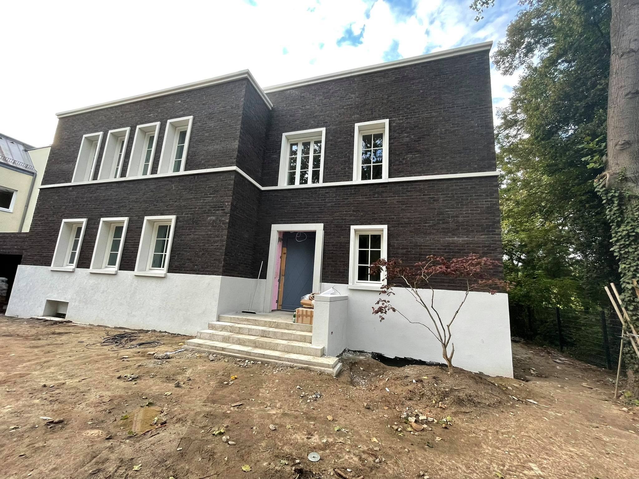 Dwukondygnacyjny dom z ciemnej cegły z białymi obramowaniami okien i gzymsami, widok z zewnątrz na częściowo zagospodarowany teren z młodym drzewkiem japońskim przed wejściem.