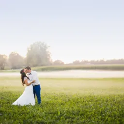 Pocałunek młodej pary na tle łąki spowitej poranną mgłą, on w niebieskich spodniach i muszce, ona w białej sukni i wianku z czerwonych kwiatów.