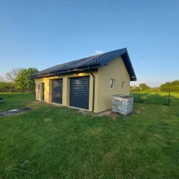 Żółty budynek garażowy z dwoma czarnymi bramami rolowanymi i czarnym dachem na tle zielonego trawnika i błękitnego nieba.