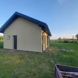 Świeżo otynkowany budynek gospodarczy z ciemnym dachem w Ludwinie, widok z zewnątrz na tle zieleni i błękitnego nieba.