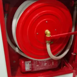 Czerwona szafka hydrantowa z rozwiniętym wężem i gaśnicą wewnątrz, zamontowana na białej ścianie.