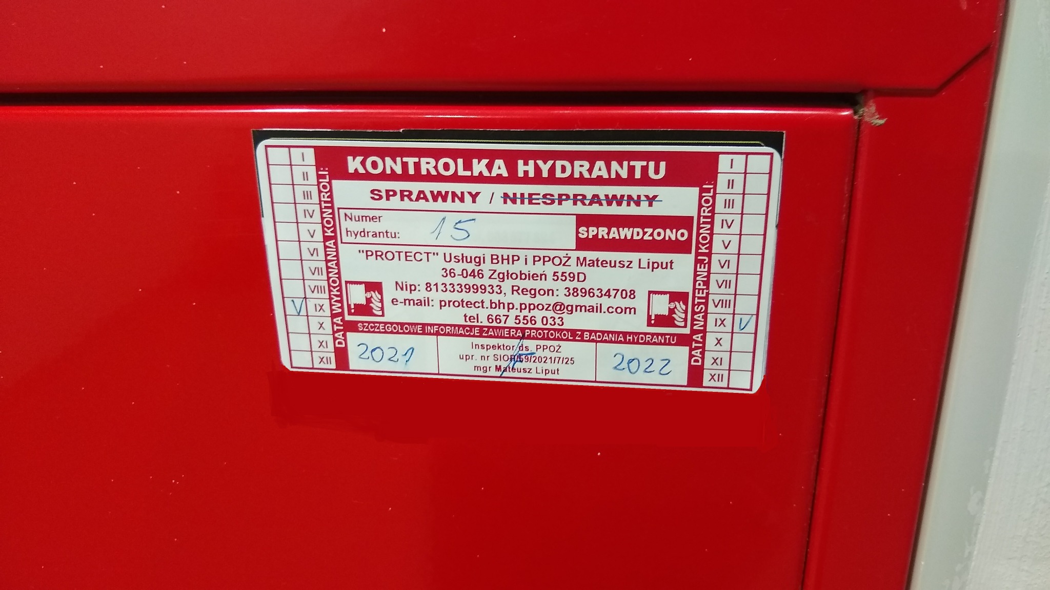 Czerwona szafka hydrantowa z naklejoną kontrolką, na której widnieje informacja o numerze hydrantu, dacie kontroli (2021) i planowanej dacie następnej kontroli (2022), a także dane firmy 'PROTECT'...