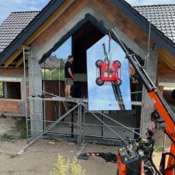 Montaż dużej aluminiowej fasady okiennej z użyciem przyssawki próżniowej mocowanej do dźwigu, widoczni pracownicy na rusztowaniu i operator dźwigu, surowy stan budynku.